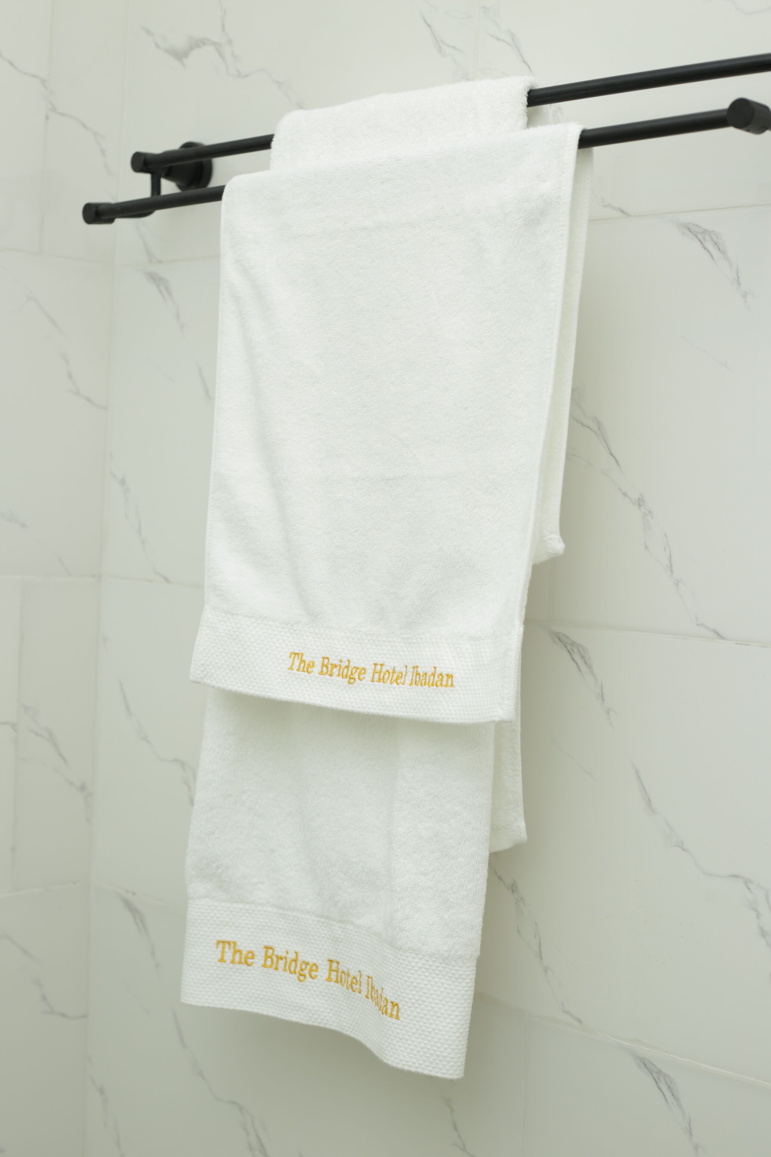 A white towel with "The Bridge Hotel Ispahan" text hanging on a black towel rack against a marble wall.