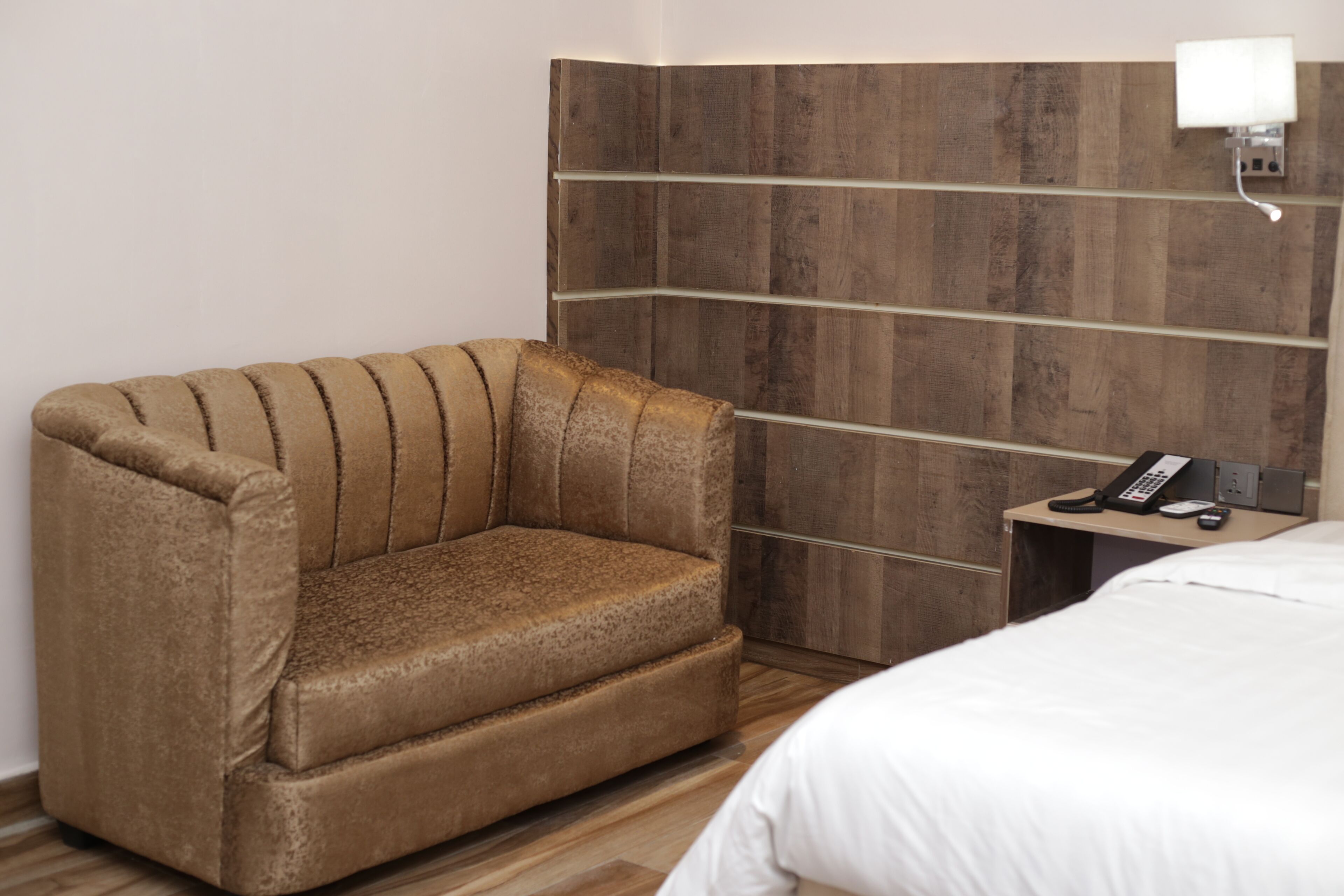 Une chambre d’hôtel avec un canapé marron, une tête de lit en bois, une table de chevet avec un téléphone et un lit blanc.