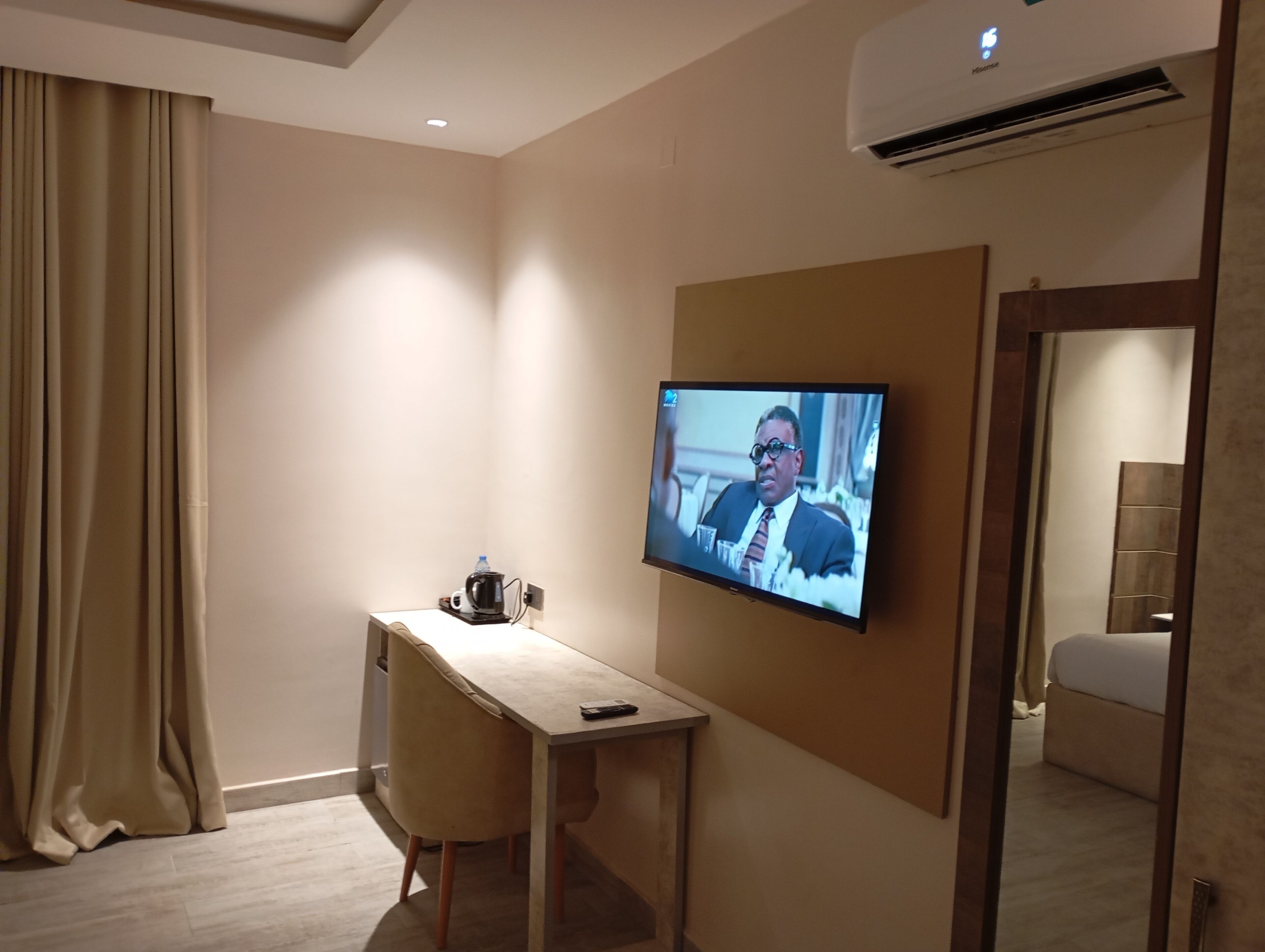 A hotel room with a flat-screen TV mounted on the wall, a wooden desk, a chair, a bed, and a large mirror.