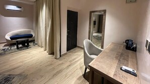 A hotel room with a wooden floor, a white armchair, a small table, a mirror, and a TV remote on the table. - The Bridge Hotel (Ibadan)