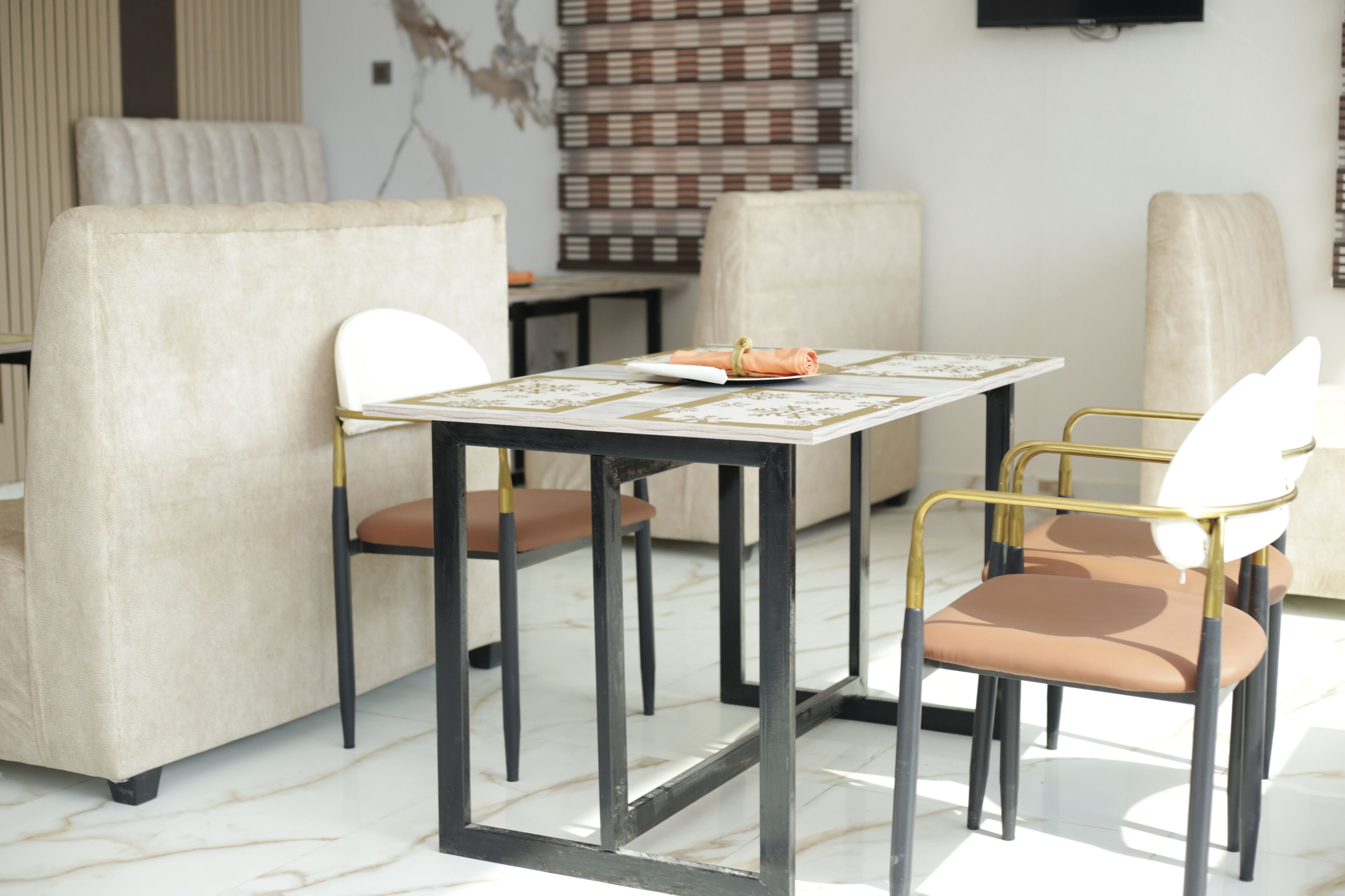 Uma área de jantar moderna com piso de mármore, uma mesa com toalha branca e dourada, e duas cadeiras com estrutura dourada e almofadas marrons.