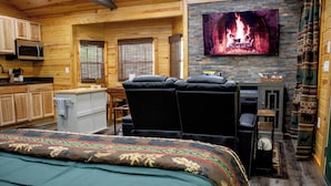 A cozy cabin interior with a bed, a TV displaying a fireplace, a kitchenette, and a seating area. - Cozy Treehouse For 2 | Outdoor Spa Tub + Fire Pit (Hot Springs)