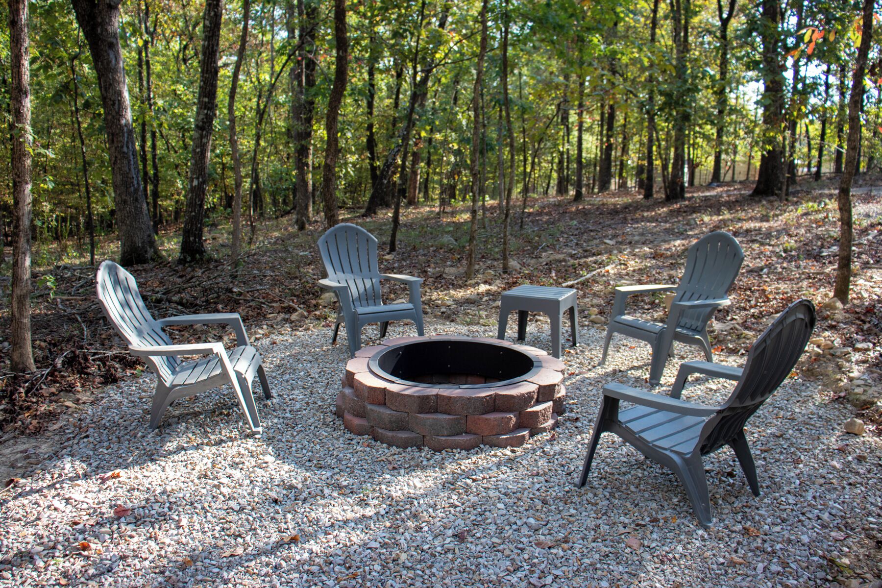 A fire pit surrounded by four Adirondack chairs in a forest setting.