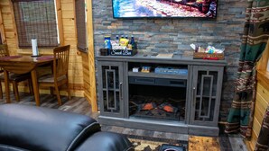 A cozy cabin interior with a wooden dining table, a leather couch, a fireplace, and a mounted TV. - Cozy Treehouse For 2 | Outdoor Spa Tub + Fire Pit (Hot Springs)