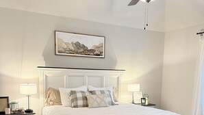 Una camera da letto con un letto bianco, due comodini, una ventola a soffitto e un quadro appeso al muro.