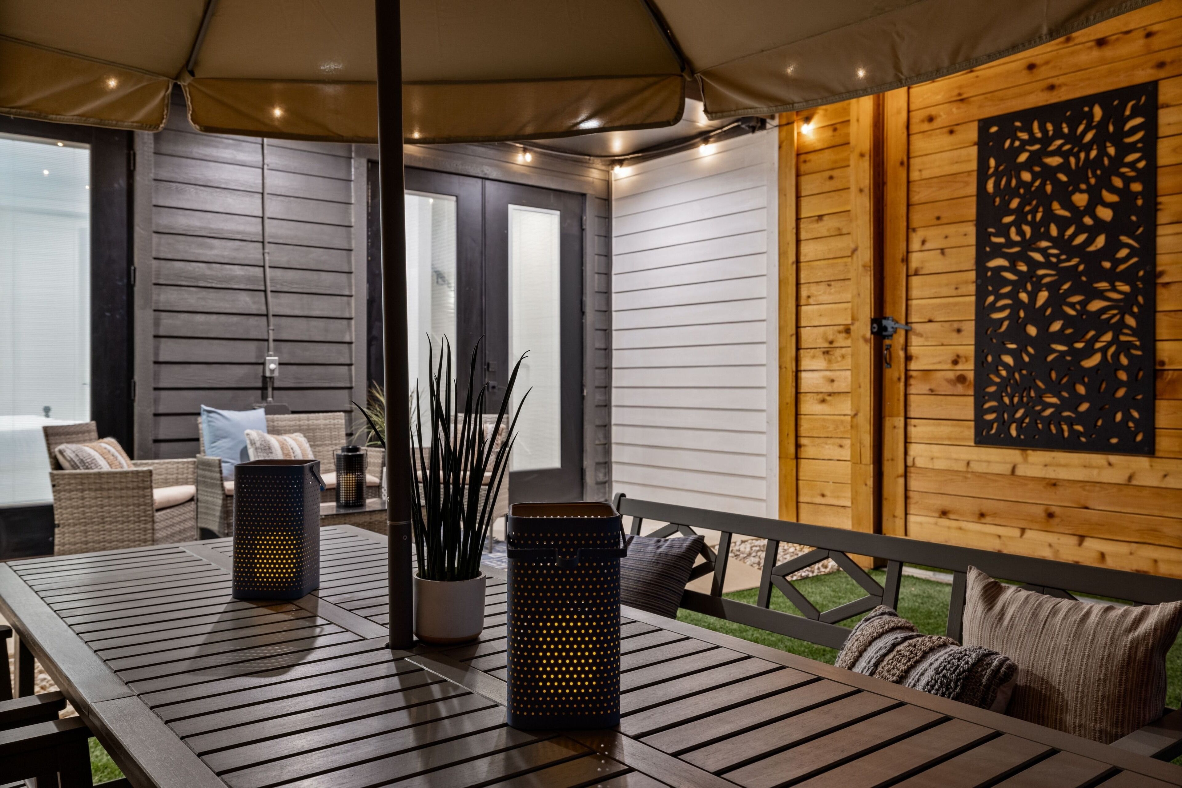 Ein überdachter Patio-Bereich mit einem Holztisch, zwei Laternen und einem Blumentopf.