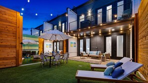 A well-lit outdoor patio area with a dining table, chairs, and a lounge chair.