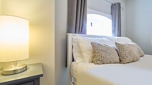 A neatly made bed with white bedding and two textured pillows, a bedside lamp on a nightstand, and a window with curtains. - Host & Stay | Roper Road (Canterbury)