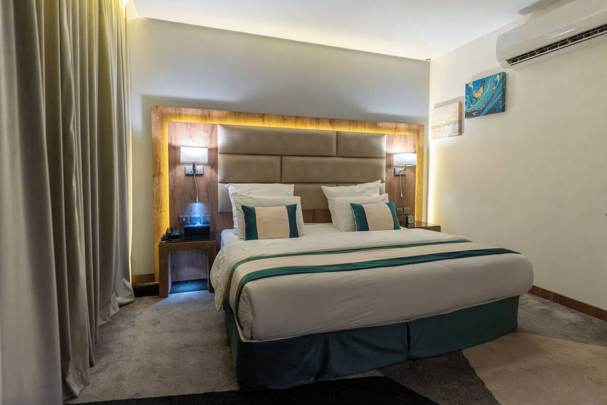 Quarto de hotel com uma cama grande, cabeceira de madeira, duas mesas de cabeceira com abajures e ar condicionado fixado na parede.