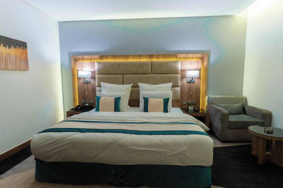 Quarto de hotel com uma cama grande, uma cadeira e uma mesinha.