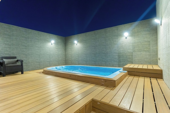 Une terrasse en bois avec un jacuzzi et une chaise, entourée de murs dotés d’un éclairage intégré.