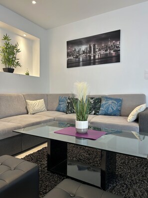 Un salon moderne avec une table basse en verre, un canapé gris et un tableau représentant un paysage urbain.