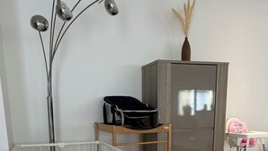 Una cuna para bebé con colchón estampado, un estante de madera, un refrigerador y una trona rosa.