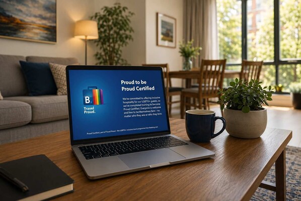 Una computadora portátil mostrando un sello Certified Proud, una planta, una taza y un libro sobre una mesa de madera en una habitación con un sofá y una lámpara.