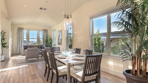 Un coin repas avec une table en bois dressée pour quatre personnes, entourée de chaises en bois foncé. Une grande fenêtre se trouve sur le côté droit, et une plante est visible au premier plan.