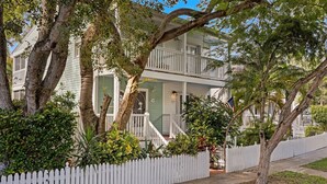 Une maison à deux étages, avec une clôture en bois peinte en blanc, entourée d’une végétation luxuriante et d’arbres.