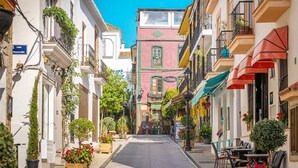Uma rua estreita, ladeada por edifícios coloridos, alguns com mesas ao ar livre e plantas.