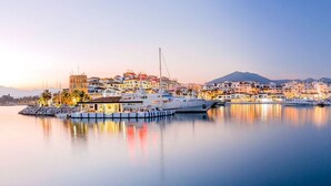 Uma marina com iates atracados, um edifício com uma torre e uma montanha ao fundo.