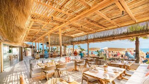 Restaurante ao ar livre com mesas e cadeiras de madeira, telhado de palha e vista para a praia.