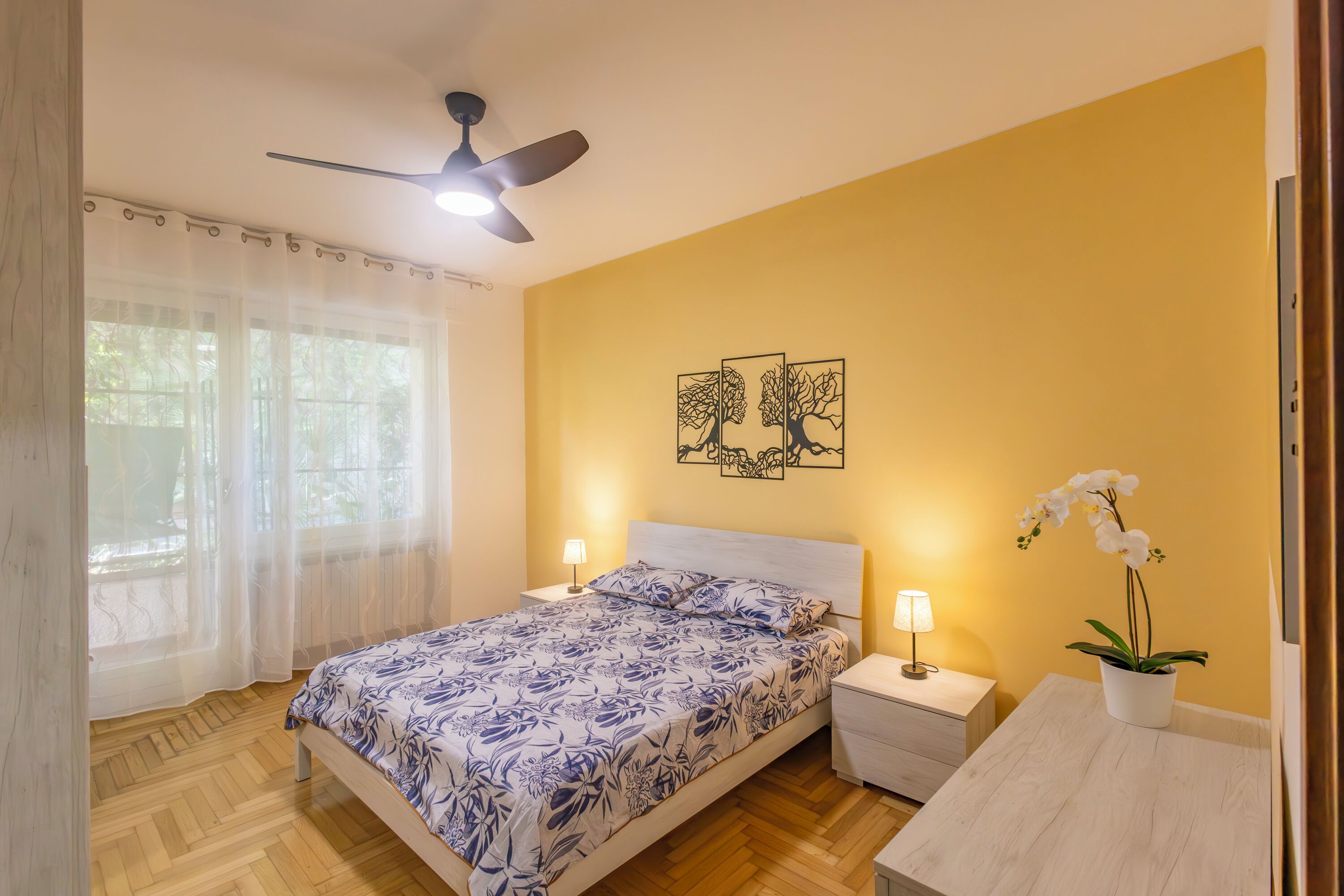 Une chambre à coucher comprenant un lit, des tables de chevet, un ventilateur de plafond, une fenêtre avec des rideaux légers et un pot d’orchidée.