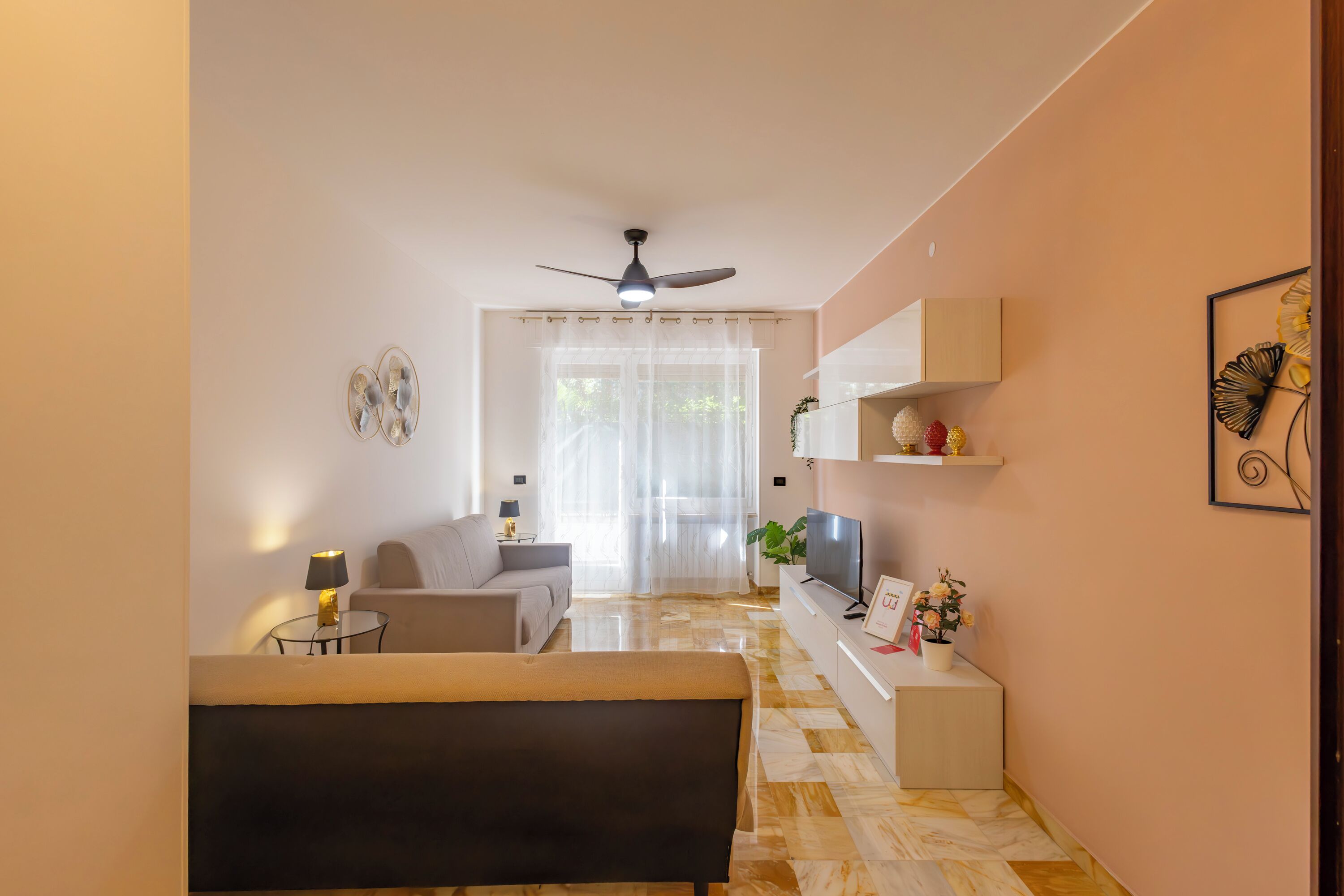 Ein modernes Wohnzimmer mit einer Couch, einem Couchtisch und einem Fernseher. Es gibt einen Deckenventilator, ein Fenster mit transparenten Vorhängen und einen Flur, der in einen anderen Raum führt.