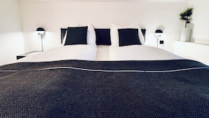 Ein ordentlich bezogenes Bett mit dunkelgrauem Überwurf und weißen Kissen, an beiden Seiten jeweils ein Nachttisch mit Lampe, in einem schlichten Zimmer mit weißen Wänden und einer kleinen Pflanze rechts.