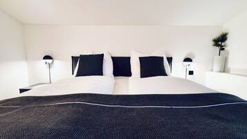 A neatly made bed with a dark bedspread and white pillows, flanked by two bedside tables with lamps, in a minimalist room with white walls and a small plant on the right.