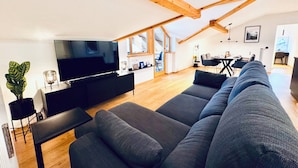 Ein modernes Wohnzimmer mit einem großen, grauen Sofa, einem Flachbildfernseher, Holzbalken an der Decke und einem Essbereich im Hintergrund.