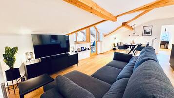 A modern living room with a large grey sofa, a flat-screen TV, wooden beams on the ceiling, and a dining area in the background.