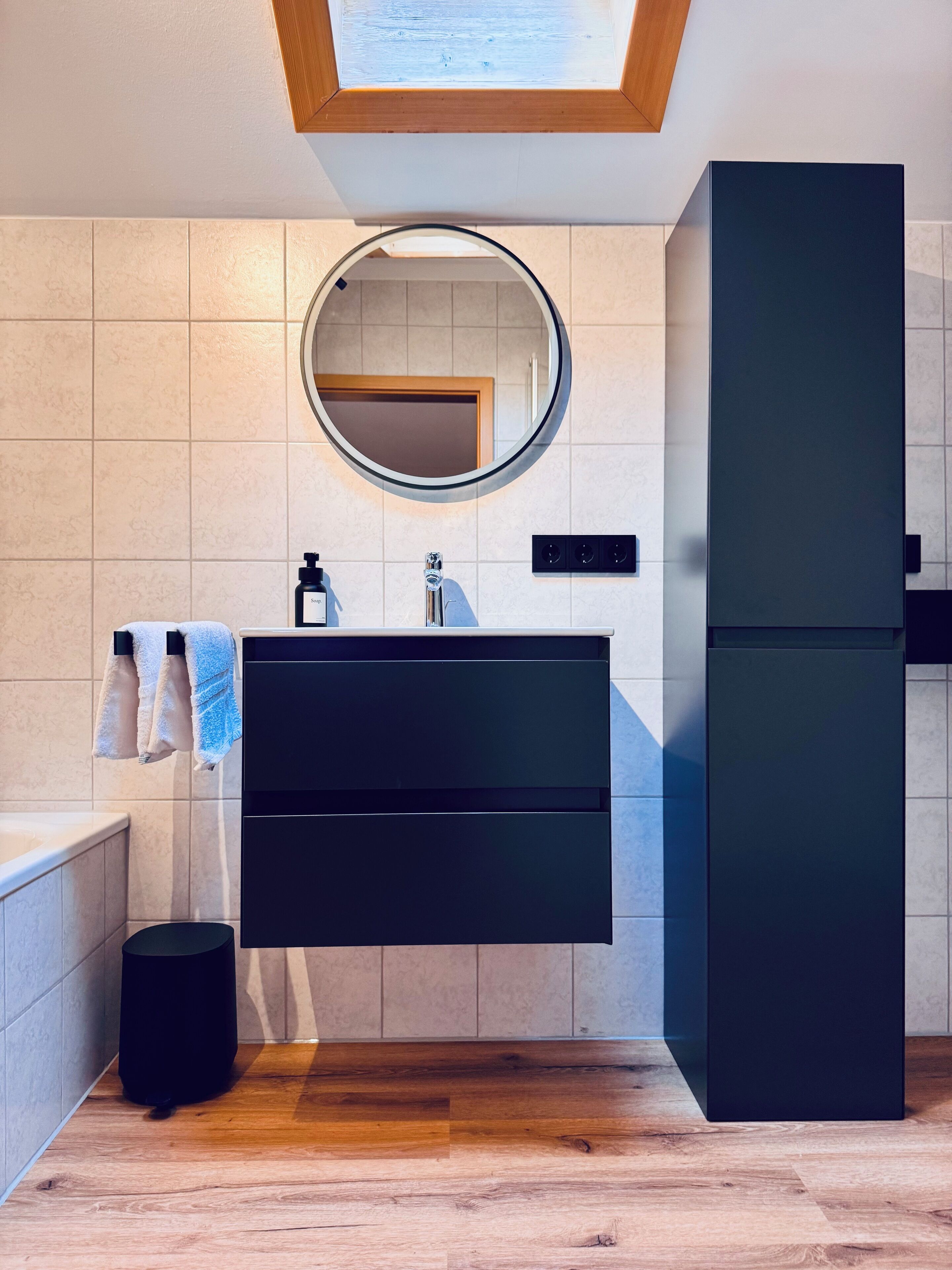 A modern bathroom with a wooden ceiling, a round mirror, a black vanity, a black trash bin, and a wooden floor.