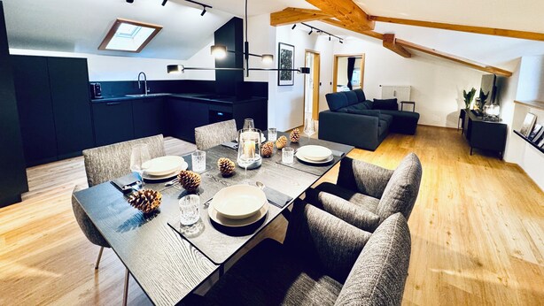 Eine moderne Küche mit dunklen Schränken, ein Essbereich mit einem Holztisch für vier Personen und ein gemütlicher Wohnbereich mit einem dunklen Sofa und einem Kamin.