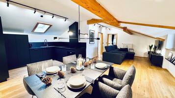 A modern kitchen with dark cabinets, a dining area with a wooden table set for four, and a cozy living space with a dark sofa and a fireplace.