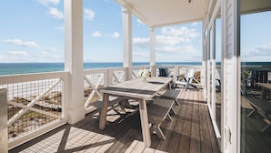 A covered outdoor deck with wooden furniture, a white railing, and a view of the ocean.