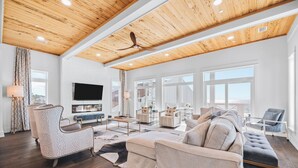 A spacious living room with a high ceiling, wooden beams, a large TV, and a view to the outside.