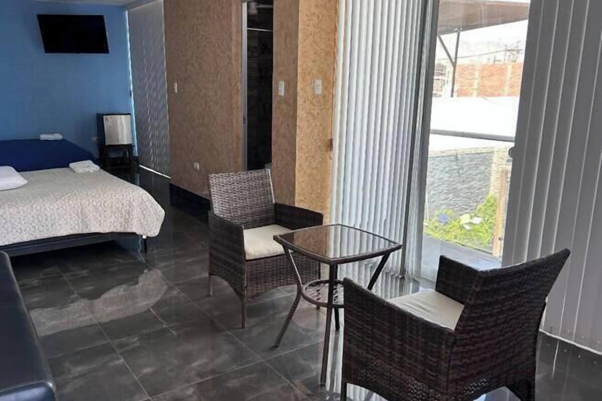 A hotel room with a bed, a small table, two wicker chairs, a TV, and a view of the outside.