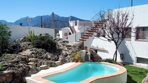 A swimming pool with a stone border, surrounded by a garden with rocks and plants, and a white building with a staircase. - Villa La Roca - A fusion of modern and typical Andalucian architecture (Montecorto)