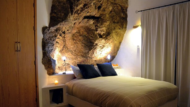 A bedroom with a large rock wall, a bed with pillows, a nightstand with a lamp, and a wooden wardrobe. - Villa La Roca - A fusion of modern and typical Andalucian architecture (Montecorto)