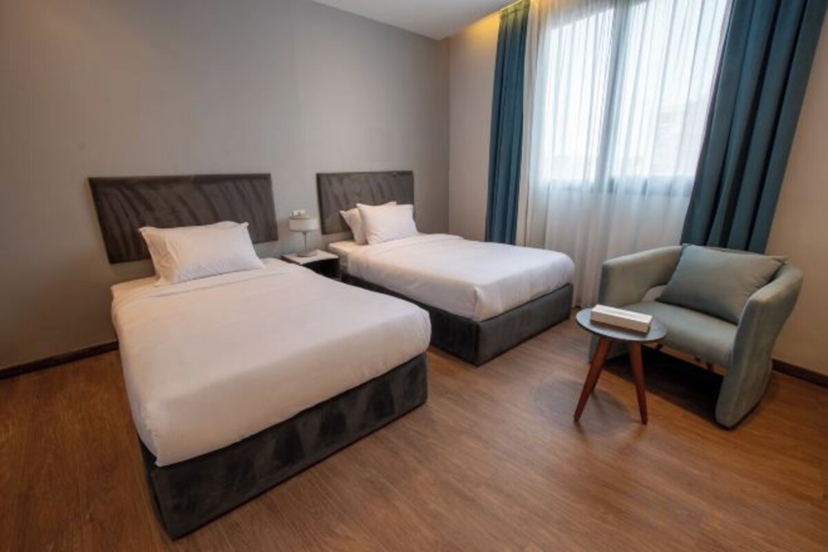 A hotel room with two beds, a small table, a chair, and a window with curtains.