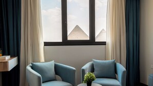 Ein Zimmer mit zwei blauen Sesseln, einem kleinen runden Tisch mit einer Pflanze und Blick auf Pyramiden durch ein Fenster mit weißen Vorhängen.