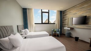 Ein Hotelzimmer mit zwei Betten, einem an der Wand montierten Fernseher, einem kleinen Tisch mit einer Pflanze und Blick auf Pyramiden durch das Fenster.