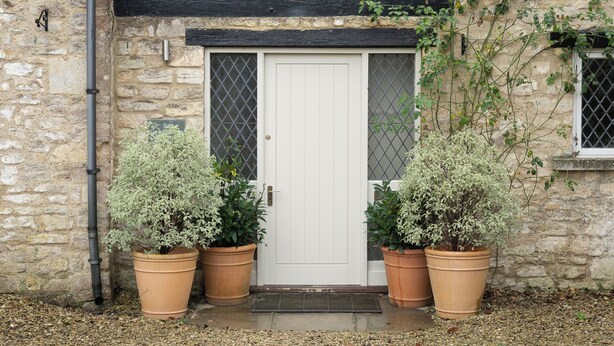 Un bâtiment en pierre avec une porte blanche, deux plantes en pot et une poutre noire au-dessus de la porte.