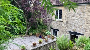 Un bâtiment en pierre doté d’un patio carrelé, entouré de différentes plantes en pot et de fougères.