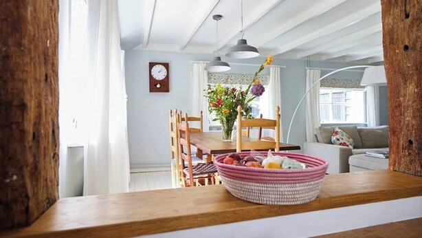 A dining area with a wooden table, chairs, a clock, and a vase with flowers. - Cottage Duinroos - Dune Rose (Oostvoorne)