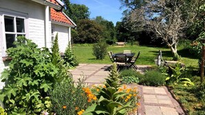A white house with a red-tiled roof, a garden with a variety of plants and flowers, a paved patio with a table and chairs, and a lawn with a bench in the background. - Cottage Duinroos - Dune Rose (Oostvoorne)