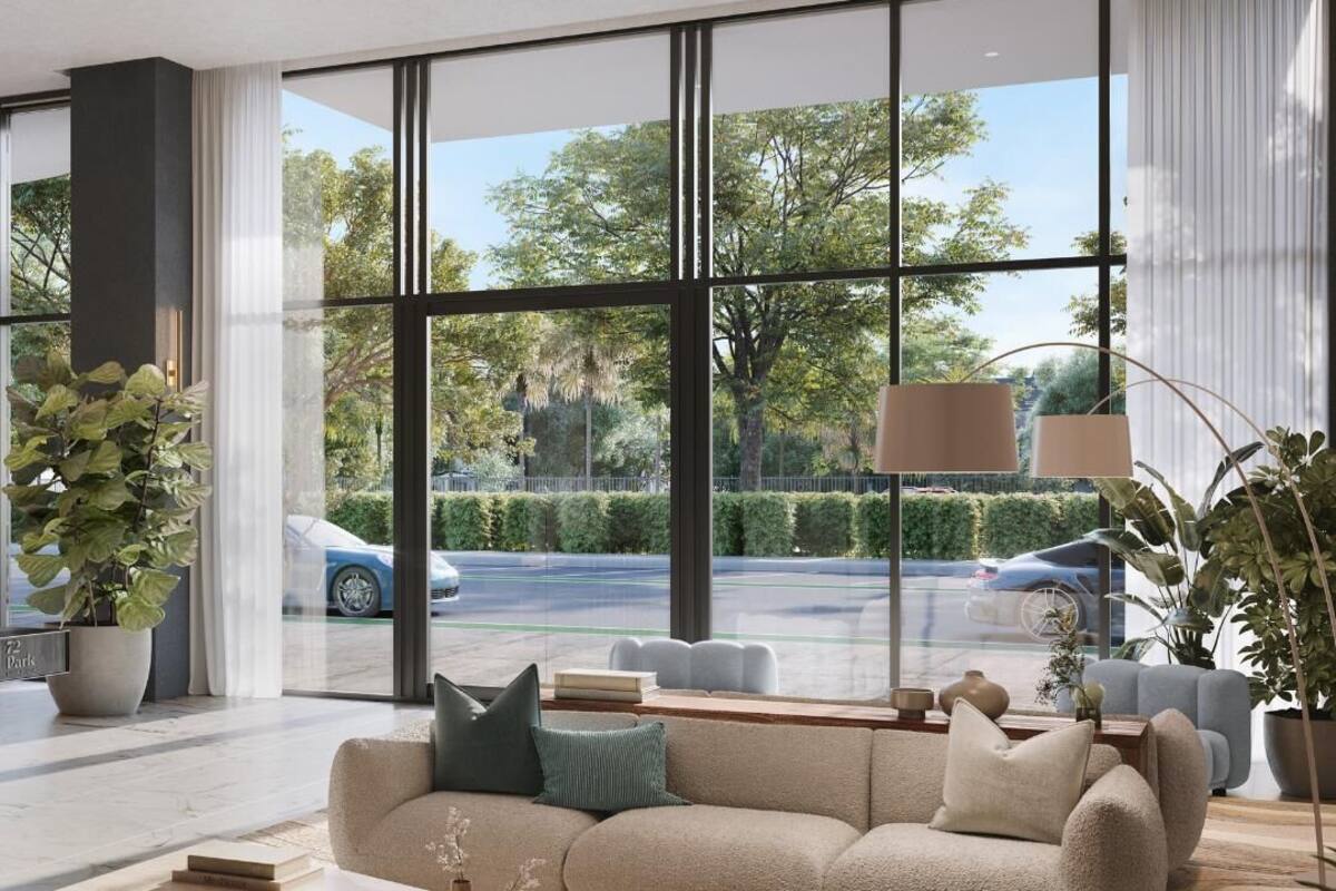 A modern living room with a beige sofa, a coffee table, and a large window offering a view of a street with cars and greenery.