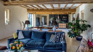 A spacious living room with a high ceiling, exposed wooden beams, a black leather sofa, a coffee table, and a potted plant.