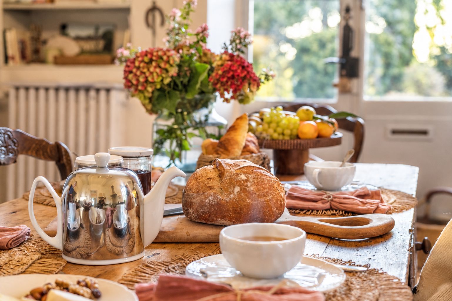 A breakfast setting with a teapot, bread, fruit, and a vase of flowers.