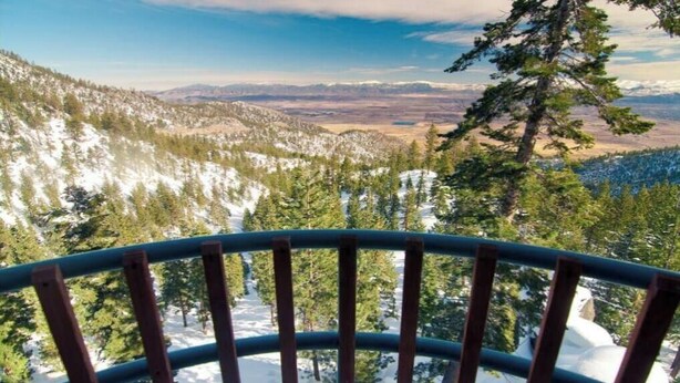 A panoramic view from a balcony with a metal railing, overlooking a snowy mountain landscape with dense forests and distant hills.