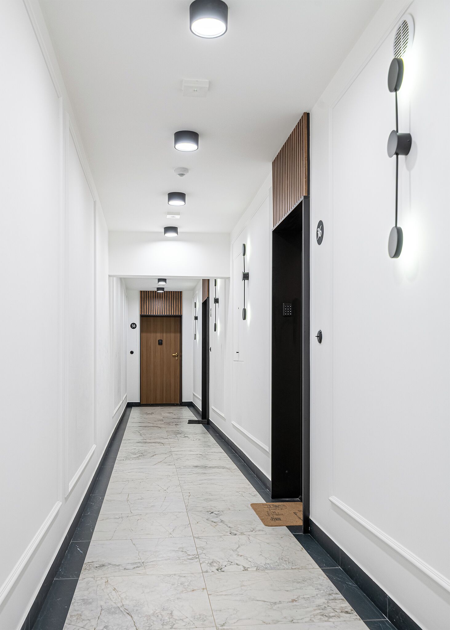 A modern hallway with white walls, black and white marble flooring, recessed lighting, and a wooden door at the end.