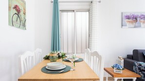 Un coin repas avec une table en bois dressée pour deux personnes, des chaises blanches, une petite table d’appoint avec une plante et une fenêtre avec des rideaux.